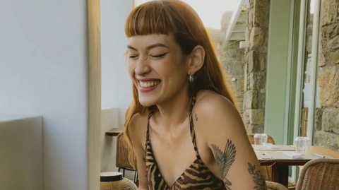 Sophie is posing in a head and shoulders shot with a wide smile, wrinkled nose and closed eyes. She has long auburn hair and a very straight, short fringe. She is sitting at a table in a restaurant conservatory and she is wearing a leopard-print strappy sundress. She has tattoos on her arm, including a bird with large outstretched wings. 