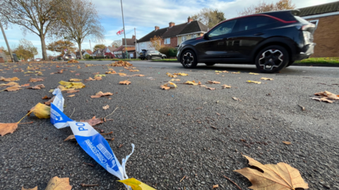 A low view of a residential road, with police tape on the ground, as well as lots of leaves. Cars and houses run across the back of the image