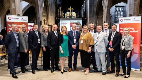 A group of people in suits and dresses standing in a cathedral in front on banners with tourism slogans on.