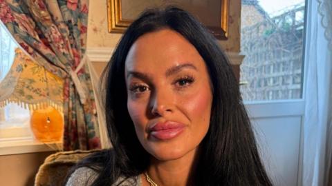 A woman with black hair and wearing a grey shirt and gold necklace. She is sitting in a living room and looking at the camera