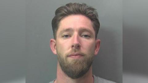 A police mugshot of Matthew Fox. He has a beard and swept-back hair and is looking straight at the camera. He is wearing a grey top and is in front of a grey background.