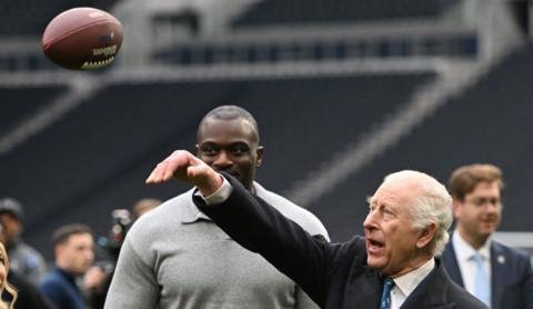 King Charles III in the act of throwing an American football into the air. He wears a smart charcoal suit, white shirt and blue tie. His arm is raised and the ball is visible in the air about a foot after he released it. Charles has his mouth open in effort. Professional American football player Efe Obada watches with a smile. Obada is tall and broadly built, wearing a grey long sleeve top.