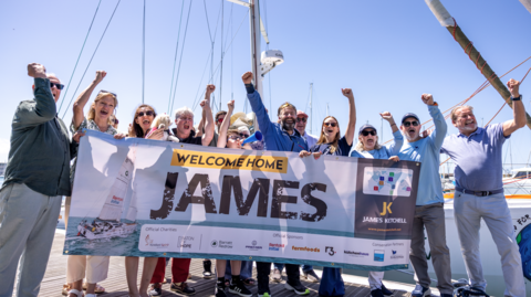 James Ketchell celebrating his return after an eleven-month voyage around the world. He is surrounded by a group of men and women who are punching the air, celebrating his return. They are holding a sign, reading "Welcome Home, James". It is a sunny day.