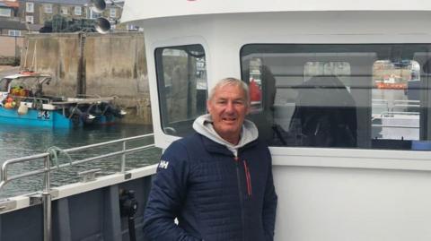 Andrew Douglas is standing by his boat in Seahouses harbour. He is a man in his 50s wearing a blue coat with a white hoodie and has short blondish grey hair.