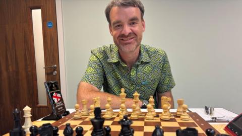 A man with greying hair wearing a short-sleeved green shirt smiles as he sits behind a wooden chess board.