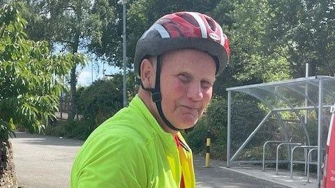 A man in a bright yellow cycling jersey and red and white helmet smiles at the camera. He is standing on quiet lane with a bike shed in the distance.