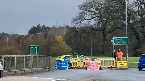 A police car at the A617 in Rainworth, Nottinghamshire. The car has green and blue markings and is behind temporary yellow plastic fencing. Three signs stand in front. A blue one on the left has "police accident" written on it in white letters. The middle one, a red one, has "road closed due to incident" also in white letters on it. The right hand one is yellow with black wording saying "Diversion" with an arrow pointing to the right. Two people stand to the right of the police car next to a sign for Newark.