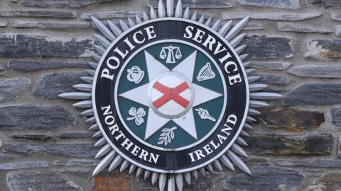 This images shows the PSNI crest - which features the St. Patrick's saltire, and six symbols representing different and shared traditions. They are the Scales of Justice, a crown, harp, a torch, an olive branch, a shamrock,