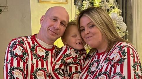 A bald man and a woman with shoulder length blonde hair are smiling at the camera, with a small child between them. All three are wearing Christmas red and white outfits