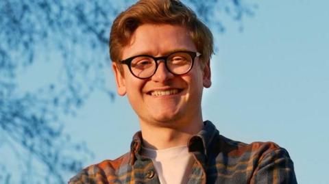 A close up of Richard. He has curly brown hair and is wearing a pair of glasses, a check shirt, and is smiling.