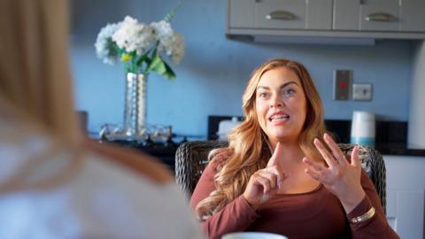 A woman with long wavy hair, sits in her pristine kitchen, mid-counting on her fingers. There are white flowers behind her.