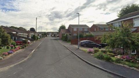A quiet residential street lined with well‑kept houses and tidy front gardens. The road gently slopes upward toward the background, where more houses can be seen. On both sides of the street, there are a mixture of detached and semi‑detached brick homes, many with driveways and parked cars. The front gardens are neatly landscaped, featuring colourful flowers, shrubs, and small trees. A few streetlights line the pavement, and the sky above is partly cloudy with patches of blue showing through.