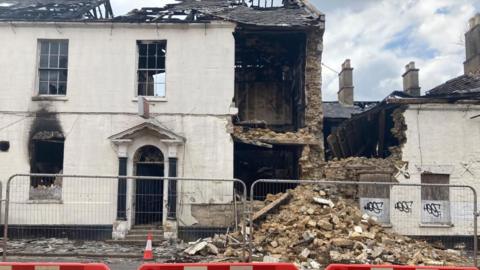 A two-storey house rendered in white. There is a Georgian door with decorative pillars either side and a pediment above. Some of the glass from the windows is missing, and there is a clear scorch mark above a ground floor window. Part of the grey roof is missing. The wall has come down on the right of the building, leaving the inside visible. There is a pile of rubble in front of the house, with metal fencing installed in front of it.