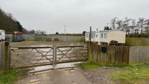 A plot of land is surrounded by a wooden fence and gate. In the right corner is a cream coloured static caravan. on the right is an old horse box and next to that is a white mobile caravan. 