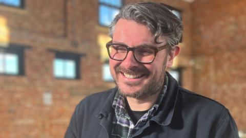Ross Millard, of The Futureheads, smiling at the camera. He is wearing a green-and-purple checked shirt and a dark blue jacket. He has short wavy hair and glasses.