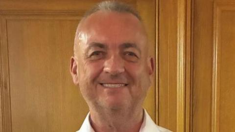 Lindsay Atkinson is wearing a white shirt and is smiling at the camera. He is standing in front of wood panelling. 