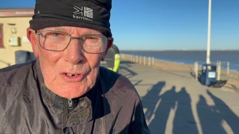 Close up image of elderly man wearing glasses and a black woolly hat on a seafront in sunshine