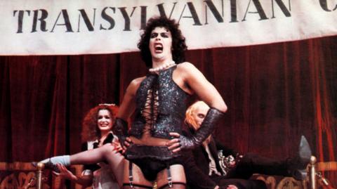 Tim Curry in a scene from the Rocky Horror Picture Show, wearing a black corset and stockings. He is singing in front of a Transylvania banner as other cast members watch