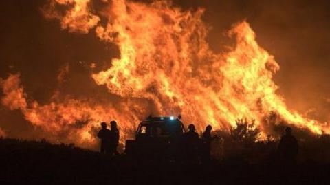 A large fire burns on a heath at night. Silhouettes of fire crews and a vehicle are visible.