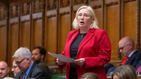 Emma Lewell MP stands giving a speech in front of the wood panelled walls of the House of Commons chamber surrounded by colleagues. She's wearing a crisp bright red blazer over a black top and has a silver crucifix necklace on. She's mid sentence holding papers in her hands in front of her. 