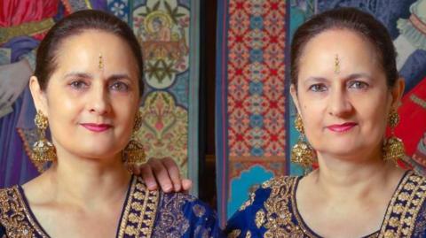 The identical twin sisters sit in front of paintings and wear cobalt blue and gold saris. One sister has her hand on her sibling's shoulder.
