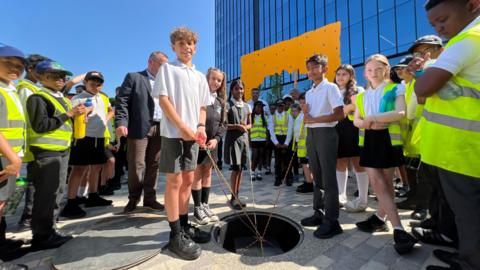 A group of 20 children stand around an open hole in the ground. Four of them are holding ropes that are crossed over each other and are around the capsule, which is a metal box that has already been lowered into the ground. In the background the blue windows of the Station Hill development are visible. 