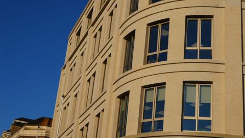 The side of a the Government of Jersey building. It has lots of windows and is a light brown colour. The sky is bright blue without a cloud.