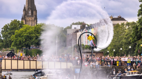A man with water jets shooting from devices attached to his feet performs a loop-the-loop on Bristol harbour while crowds watch from the quayside and a nearby bridge