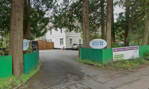 Picture shows the driveway and entrance to the hospice. There is a green fence visible, with Julia's House branding and signage attached to the fence and the entrance
