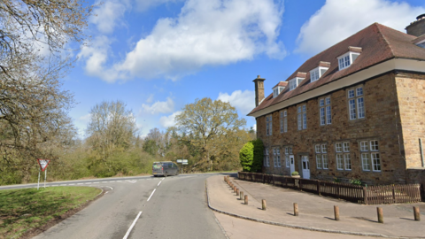 A 17th Century hunting lodge hotel stands at a T-junction in the Forest of Dean on a sunny day. A van can be seen turning right. The junction is surrounded by trees.