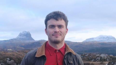 Matthew is wearing a red shirt and brown jacket with a wide collar. Behind him are mountains and sky.