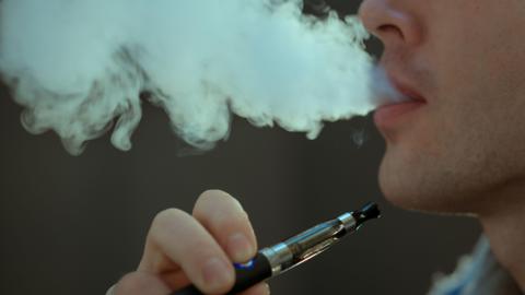 A close-in picture of a young man smoking a vape and blowing out a cloud of light grey smoke