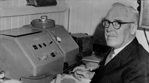 Black and white image of Sir Ernest Marsden, who is wearing a pair of spectacles, looking at the camera as he poses while sitting by a scientific machine.