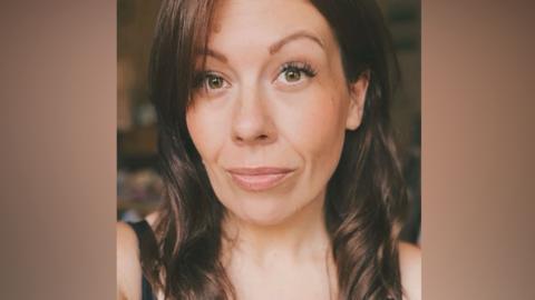 A woman with dark brown hair looks directly at the camera with green/brown eyes, a smile and a small nose ring.