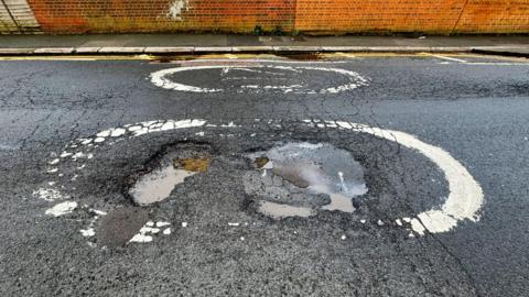 The image shows a pothole in the middle of a road, with some dirty water in them.