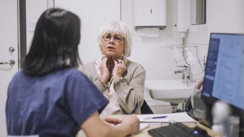 older female patient sits opposite female doctor, describing her symptoms of neck pain