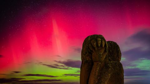 The northern lights have created a striking line of pink across the sky. Starlight is poking though. There is a moss-covered grey statue in the foreground.