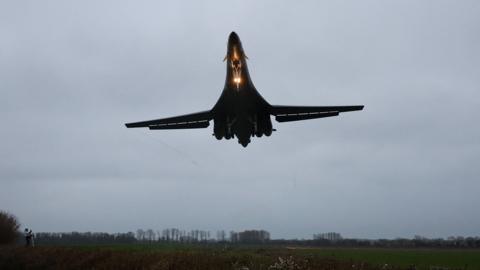 A large bomber aircraft is pictured with a bright light shining as it approaches a runway at an airbase.
