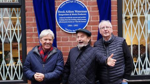 Mike Stock, Matt Aitken and Pete Waterman standing under their Historic England blue plaque outside Vine Yard Studios. Mike Stock is to the left, wearing a blue jacket and red scarf, smiling at the camera, Matt Aiken is in the middle with his left arm out and large a grin on his face. He is wearing a blue coat and flat cap and Pete Waterman is wearing a navy gilet and glasses and smiling at the camera. 