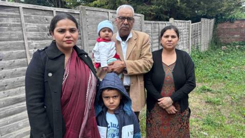 Five people, including two children, stand together outdoors by a wooden fence.
