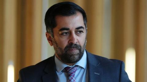 Humza Yousaf looking off camera. He has dark hair and a dark beard with grey hairs in it. He is wearing a dark suit over a light shirt with a multi-coloured tie.