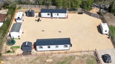 An aerial photograph of a piece of land with a number of buildings on it, caravans and mobile homes. There is grass, and laid track, also a trampoline. A fence is all around the enclosure.