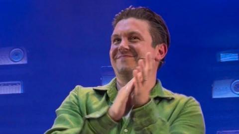 A man in a green shirt grinning and applauding, with a small headset microphone visible.