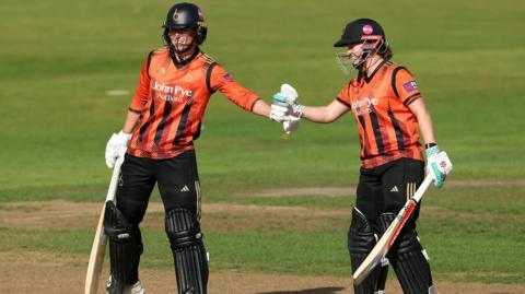 Georgia Elwiss and Kathryn Bryce meet in the middle of the crease to fistpump after a boundary, both are looking onwards. They are both wearing the orange metro bank 2025 Blaze kit. 