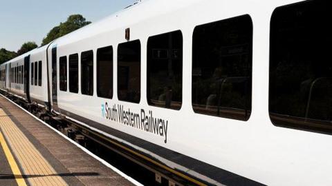 A South Western Railway passenger train stands at a platform. There are yellow markings on the platform and trees in the background under a blue sky.