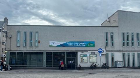 A grey rectangular building in Inverness city centre. There are two levels and long thing windows. The entrance to the building is glass. There are bikes, bins and pedestrians on the pavement outside. It's a cloudy sky. 