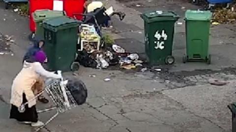 A woman and child dumping rubbish next to a number of green bins and other rubbish