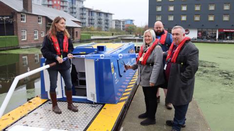 Two men and a woman stand next to a canal boat wearing red life jackets. The woman is holding a pair of scissors. On the canal is a woman holding the boat's tiller.  A blue ribbon is stretched out between the woman and one of the men as the other woman is about to cut it. The boat is sitting on the water, mostly covered in green, with buildings visible in the background.