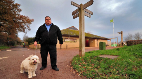 Richard Udall Worcester Woods. He is holding a small white fluffy dog on a lead. He is wearing a blue shirt underneath a black jacket and black trousers.