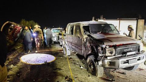 A car, blackened from a bomb explosion, is parked on a dirt road and surrounded by people.